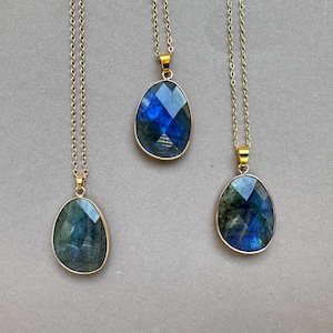 May include: Three gold-toned necklaces with labradorite pendants. Each pendant has a faceted, oval-shaped labradorite stone with blue and green iridescence, set in a gold bezel. The necklaces hang on delicate gold chains.