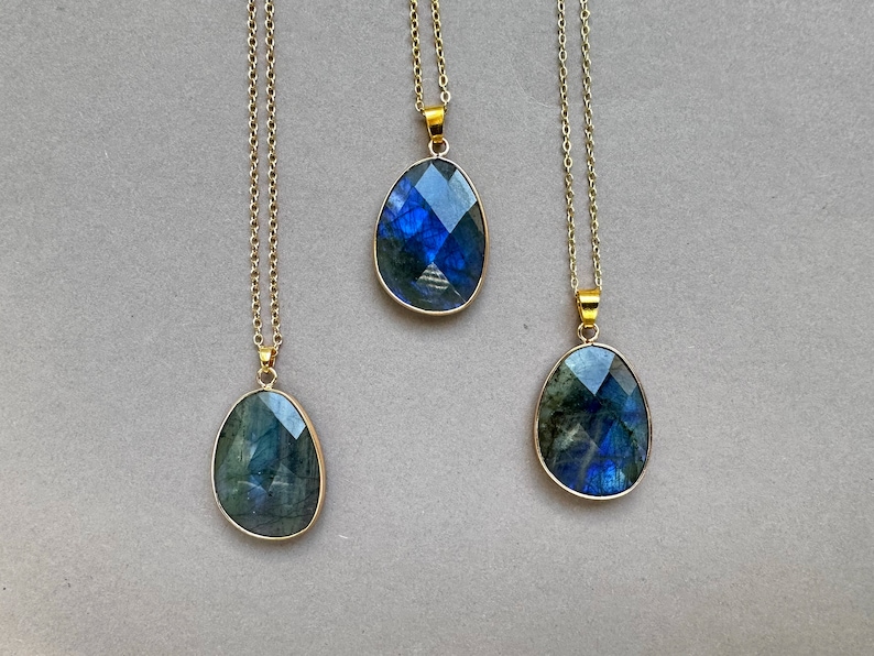 May include: Three gold-toned necklaces with labradorite pendants. Each pendant has a faceted, oval-shaped labradorite stone with blue and green iridescence, set in a gold bezel. The necklaces hang on delicate gold chains.