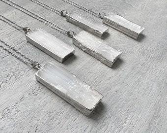 May include: Five rectangular selenite crystal pendants on silver-tone chains. The pendants have a natural, textured appearance and are displayed on a gray surface. The necklaces are a minimalist style.