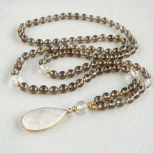 May include: A brown smoky quartz mala necklace with a clear quartz pendant. The necklace is made with 6mm beads and has a gold accent.
