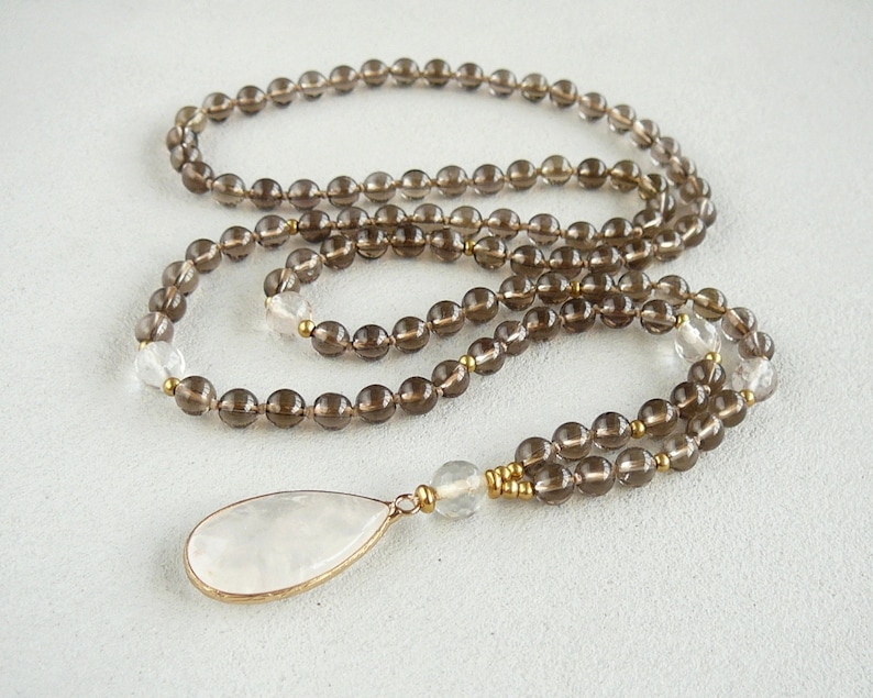 May include: A brown smoky quartz mala necklace with a clear quartz pendant. The necklace is made with 6mm beads and has a gold accent.