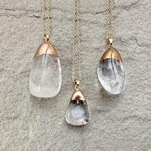 May include: Three necklaces featuring clear, teardrop-shaped pendants with gold-toned metal caps. Each pendant hangs from a delicate gold-toned chain. The stones exhibit natural internal inclusions and variations in shape, suspended from simple metal bails.