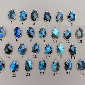 May include: A collection of 26 teardrop-shaped labradorite gemstone pendants with silver-colored metal settings. Each pendant has a unique pattern of blue, teal, and gray hues. The pendants are numbered 1 through 26.