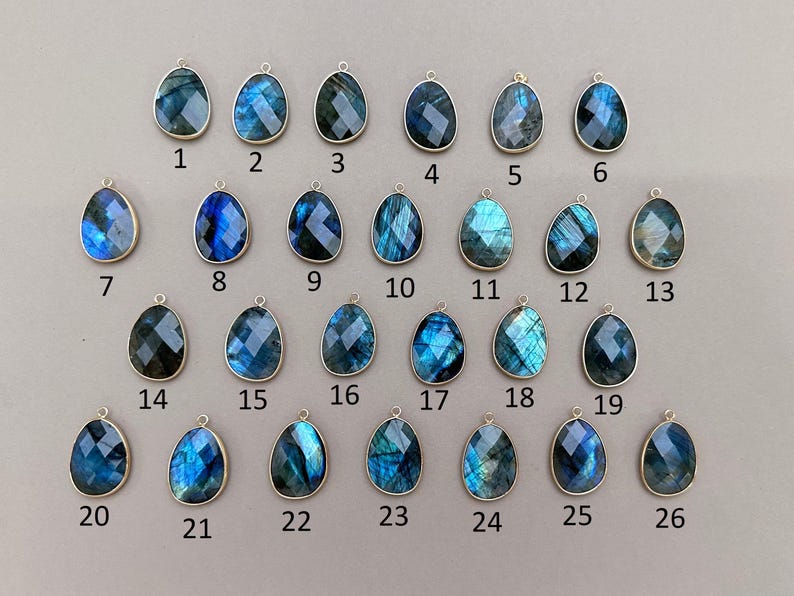 May include: A collection of 26 teardrop-shaped labradorite gemstone pendants with silver-colored metal settings. Each pendant has a unique pattern of blue, teal, and gray hues. The pendants are numbered 1 through 26.