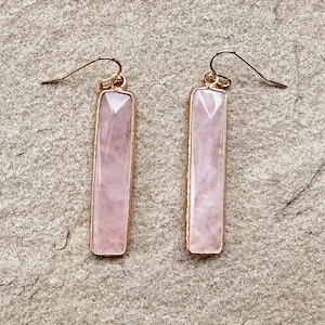 Könnte beinhalten: Ein Paar Ohrringe aus Rosenquarz mit goldfarbenen Metallakzenten. Die Ohrringe sind rechteckig und haben ein facettierten Design.