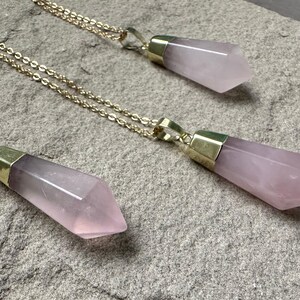 May include: Three gold necklaces with pink crystal pendants. Each pendant is a long, pointed shape with a gold cap at the top.