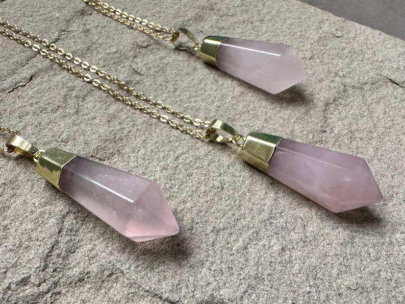 May include: Three gold necklaces with pink crystal pendants. Each pendant is a long, pointed shape with a gold cap at the top.