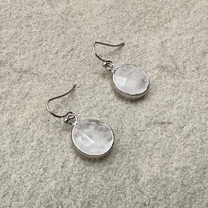 Könnte beinhalten: Ein Paar silberfarbene Ohrhänger. Jeder Ohrring hat einen runden, facettierten, weißen Edelstein, der in eine silberne Lünette gefasst ist. Die Ohrringe haben einen einfachen Hakenverschluss.