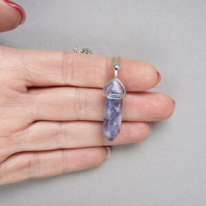 Puede incluir: Un collar con colgante que presenta un cristal de lepidolita morado y blanco, tallado en forma hexagonal de doble terminaci&oacute;n. La piedra est&aacute; asegurada en un engaste de metal plateado y cuelga de una fina cadena de eslabones plateados.