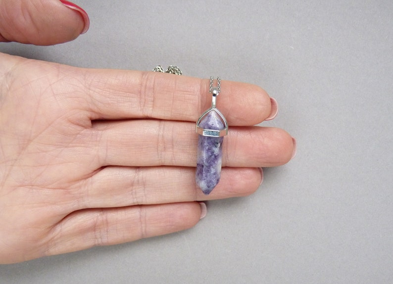 Puede incluir: Un collar con colgante que presenta un cristal de lepidolita morado y blanco, tallado en forma hexagonal de doble terminaci&oacute;n. La piedra est&aacute; asegurada en un engaste de metal plateado y cuelga de una fina cadena de eslabones plateados.
