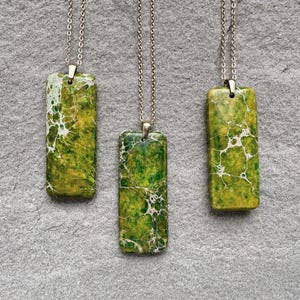 Puede incluir: Tres collares con colgantes rectangulares y cadenas plateadas. Los colgantes son una mezcla de verde y amarillo con vetas blancas. Los collares se exhiben sobre una superficie gris texturizada.