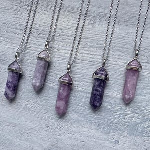 Puede incluir: Cinco collares con colgantes de punta hexagonal hechos de piedra morada y lavanda, cada uno rematado con una anilla de metal plateado y suspendido de una fina cadena plateada. Las piedras var&iacute;an en tono desde lila claro hasta violeta intenso.