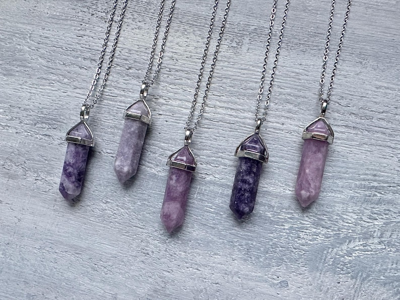 Puede incluir: Cinco collares con colgantes de punta hexagonal hechos de piedra morada y lavanda, cada uno rematado con una anilla de metal plateado y suspendido de una fina cadena plateada. Las piedras var&iacute;an en tono desde lila claro hasta violeta intenso.