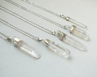 May include: Several clear quartz crystal necklaces with silver-colored chains and caps. The crystals are elongated and vary slightly in size and shape. The necklaces are displayed on a light gray surface.