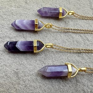 May include: Four necklaces featuring pointed, hexagonal amethyst crystal pendants in varying shades of purple and white, each secured in a gold-toned metal cap and attached to a delicate gold-toned chain, arranged horizontally in a vertical row.
