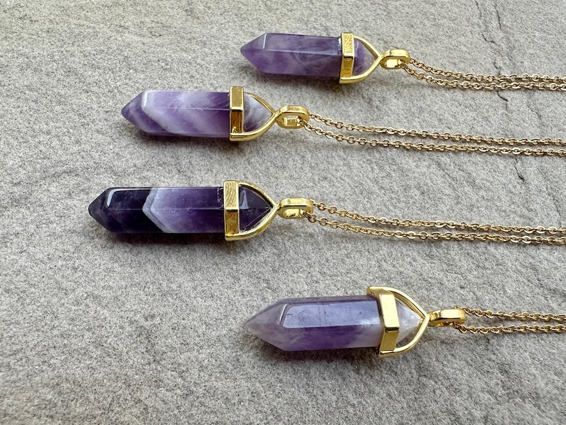 May include: Four necklaces featuring pointed, hexagonal amethyst crystal pendants in varying shades of purple and white, each secured in a gold-toned metal cap and attached to a delicate gold-toned chain, arranged horizontally in a vertical row.
