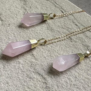May include: Three gold necklaces with pink crystal point pendants. The necklaces are layered and the crystal points are different sizes.