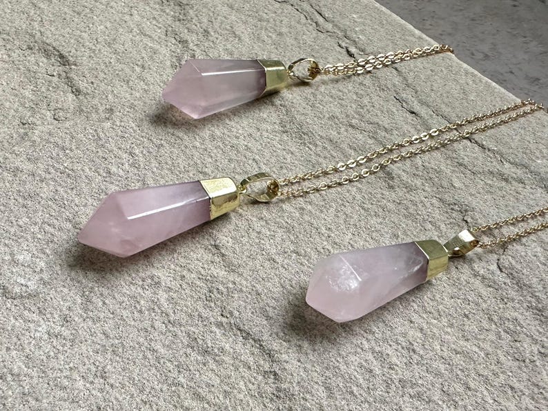 May include: Three gold necklaces with pink crystal point pendants. The necklaces are layered and the crystal points are different sizes.