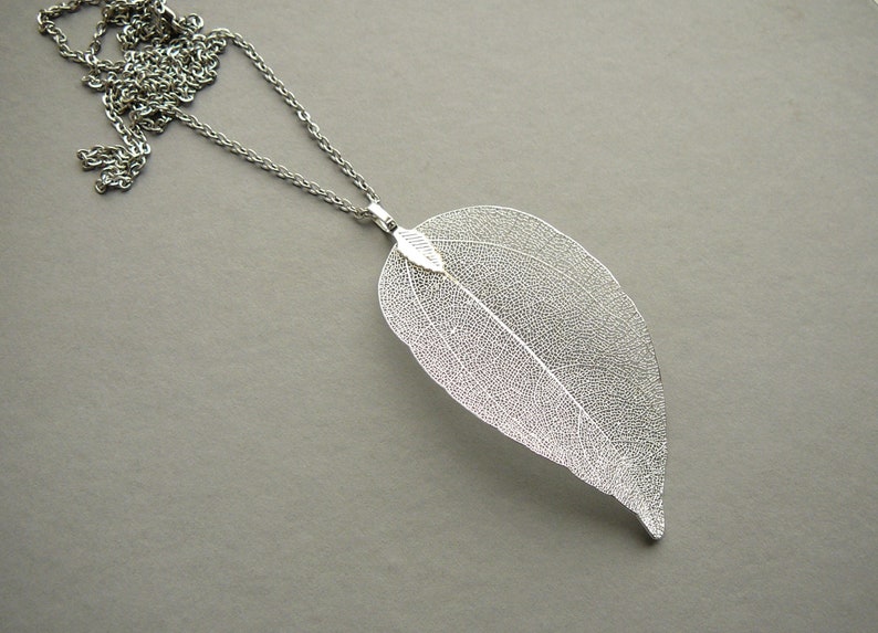 Op de afbeelding: Een zilveren ketting met een delicate bladeren hanger. Het blad is gemaakt van een dun, doorschijnend materiaal en heeft een gedetailleerd adernetwerk.