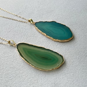 May include: Two necklaces featuring irregular, flat agate slices suspended from thin gold-tone chains. One pendant is a deep teal, while the other is a lighter olive green. Both stones have natural banding patterns and are edged in a metallic gold finish.