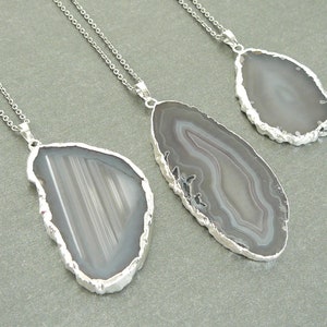 May include: Three silver necklaces with agate pendant charms. The pendants are different shapes and sizes, with varying patterns of gray and white agate.