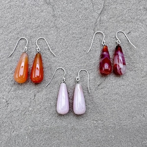 Puede incluir: Tres pares de pendientes colgantes con piedras en forma de lágrima. Los pendientes son de plata y presentan piedras naranja, rosa y roja.