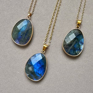 May include: Three labradorite gemstone pendants with gold-colored chains. The teardrop-shaped stones have a faceted cut, displaying iridescent blues and greens. The pendants are set in gold-colored bezels.