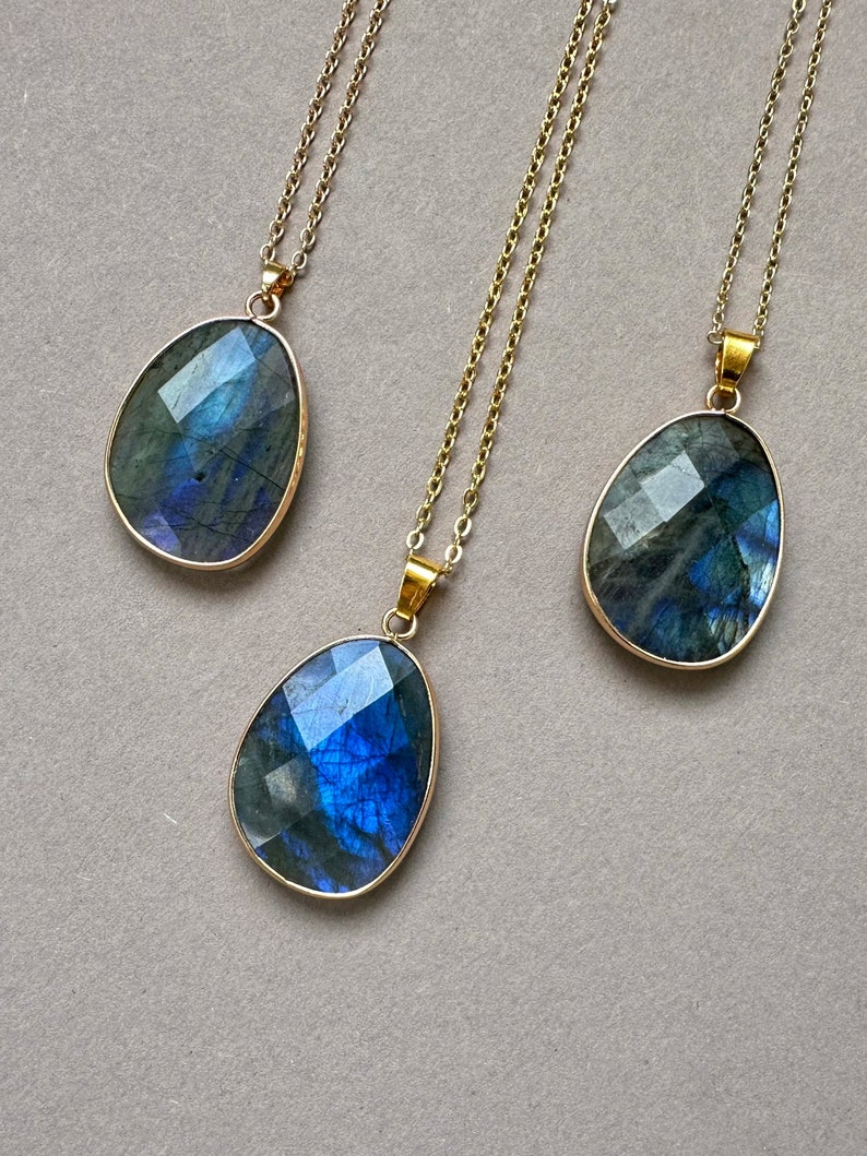May include: Three labradorite gemstone pendants with gold-colored chains. The teardrop-shaped stones have a faceted cut, displaying iridescent blues and greens. The pendants are set in gold-colored bezels.
