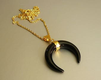 May include: A gold-colored chain necklace with a black crescent-shaped pendant. The pendant is accented with gold-colored metal at the top. The necklace is displayed on a light gray surface.