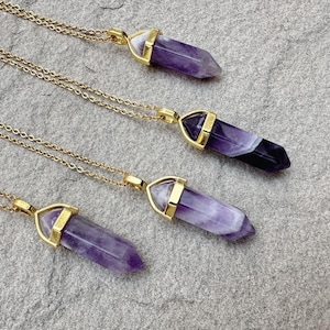 May include: Four necklaces featuring polished, hexagonal amethyst crystal points suspended from thin gold-toned metal chains. Each crystal displays natural variations in purple and white banding, secured by a gold-toned metal cap at the top.