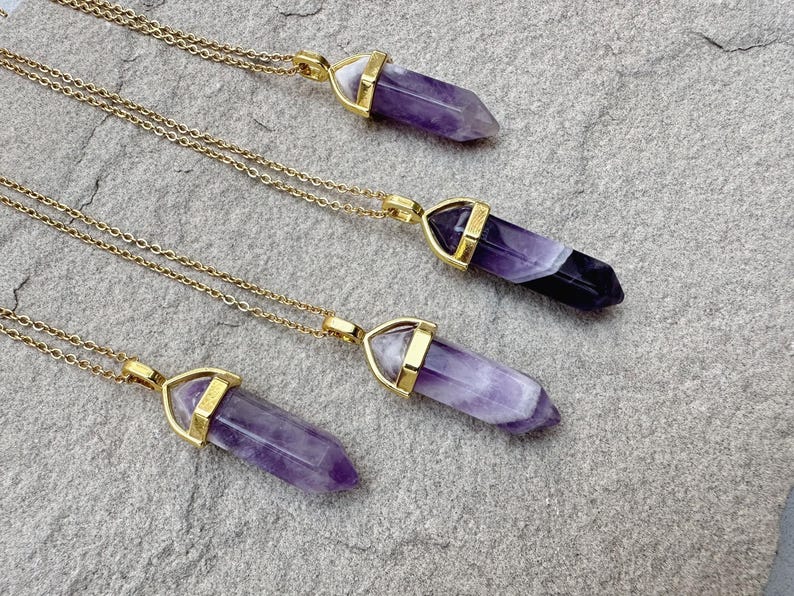 May include: Four necklaces featuring polished, hexagonal amethyst crystal points suspended from thin gold-toned metal chains. Each crystal displays natural variations in purple and white banding, secured by a gold-toned metal cap at the top.