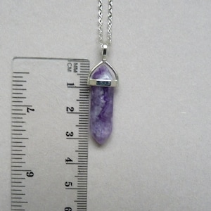 Puede incluir: Un collar de tono plateado con un colgante de cristal morado de forma hexagonal y terminaci&oacute;n en punta. Una regla de pl&aacute;stico transparente est&aacute; colocada junto al colgante, mostrando que el cristal mide aproximadamente 3,8 cm de largo.