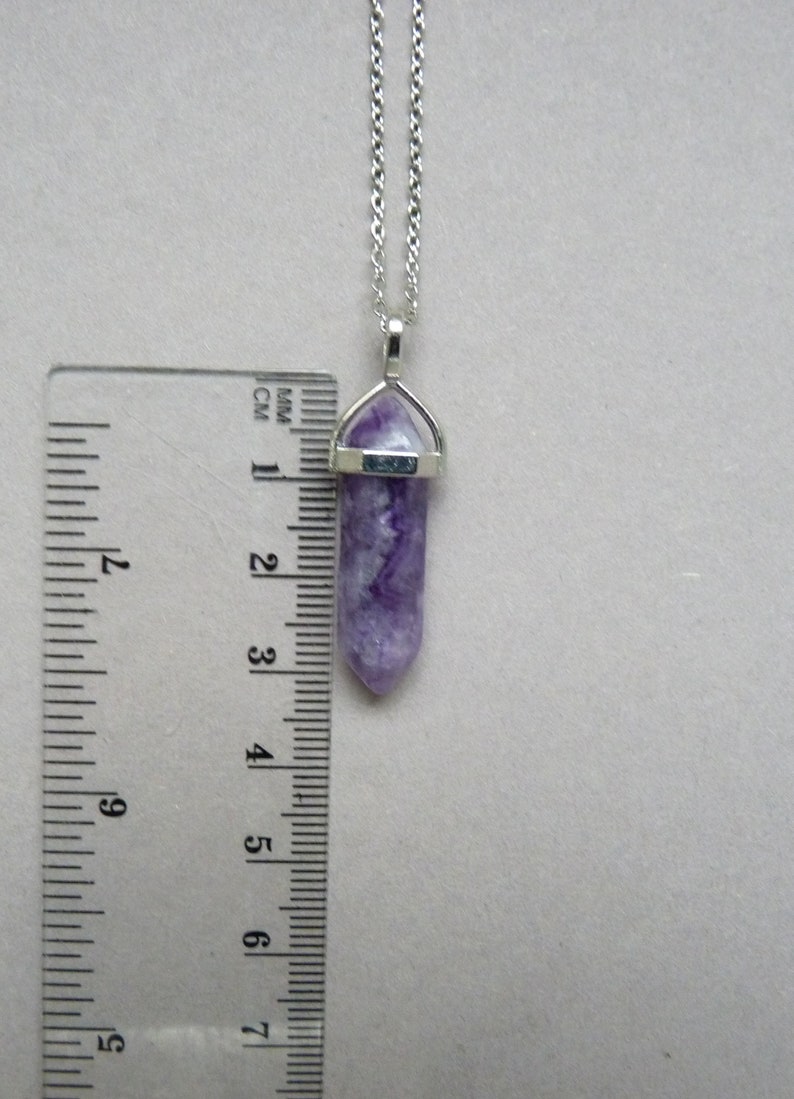Puede incluir: Un collar de tono plateado con un colgante de cristal morado de forma hexagonal y terminaci&oacute;n en punta. Una regla de pl&aacute;stico transparente est&aacute; colocada junto al colgante, mostrando que el cristal mide aproximadamente 3,8 cm de largo.