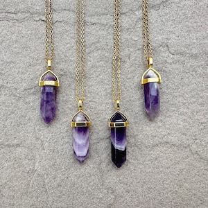 May include: Four necklaces featuring pointed, hexagonal purple amethyst crystal pendants suspended from thin gold-toned metal chains. Each crystal is secured by a gold-toned metal cap at the top, showcasing varying shades of deep violet and white banding.