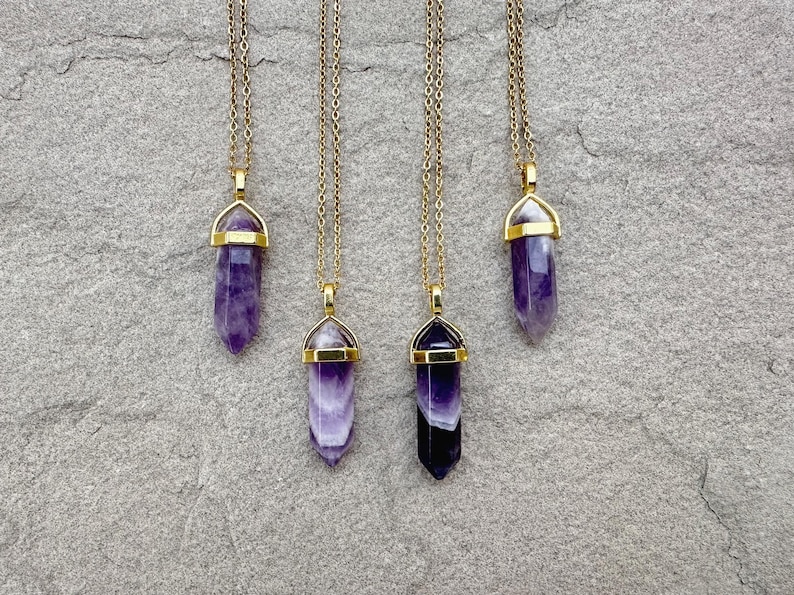 May include: Four necklaces featuring pointed, hexagonal purple amethyst crystal pendants suspended from thin gold-toned metal chains. Each crystal is secured by a gold-toned metal cap at the top, showcasing varying shades of deep violet and white banding.