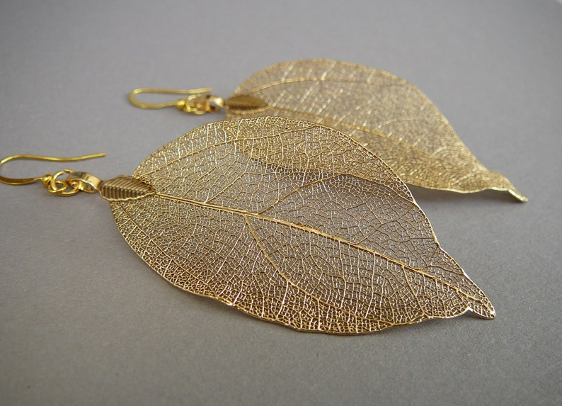 May include: A pair of gold-colored leaf earrings. The earrings are shaped like large leaves with intricate vein detailing. The earrings hang from gold-colored ear wires. The background is a solid gray.