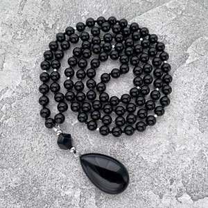 May include: A black onyx mala necklace with a teardrop-shaped pendant. The necklace is made of round beads and is approximately 54 inches long.