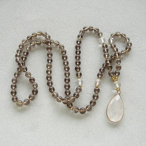 May include: A long brown gemstone necklace with a clear teardrop pendant. The necklace is made of small round beads and has a gold accent.