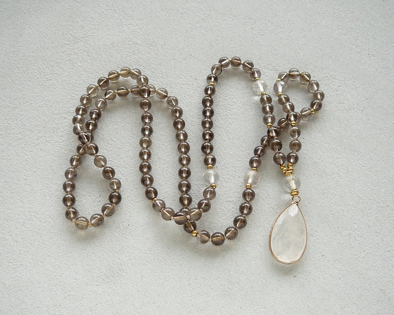 May include: A long brown gemstone necklace with a clear teardrop pendant. The necklace is made of small round beads and has a gold accent.