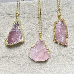 Pode incluir: Três colares dourados com pingentes de quartzo rosa bruto, rosa claro. O quartzo rosa tem formato irregular e bordas douradas. Os colares são exibidos em uma superfície texturizada, cinza claro.