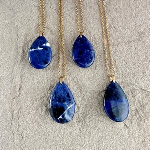 May include: Four gold necklaces with blue and white teardrop-shaped gemstone pendants. The necklaces are on a grey textured surface.