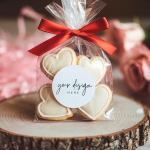 May include: A clear plastic bag filled with heart-shaped cookies tied with a red ribbon. The bag has a white circle sticker with the text "your design here". The cookies are on a wooden slice.