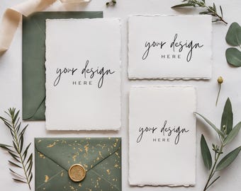May include: A flat lay featuring stationery elements. Includes four white textured cards with the text "your design HERE", two green envelopes, a gold wax seal, and sprigs of greenery. A cream-colored ribbon is also present.