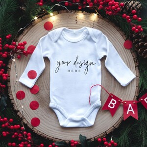 Op de afbeelding: Witte baby romper met lange mouwen met de tekst "your design here" op een achtergrond van een houten schijf, omringd door rode bessen, dennentakken en een rode "baby"-banner.