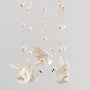 Puede incluir: Un m&oacute;vil de madera blanco con decoraciones de cisne de fieltro y pompones blancos colgando.
