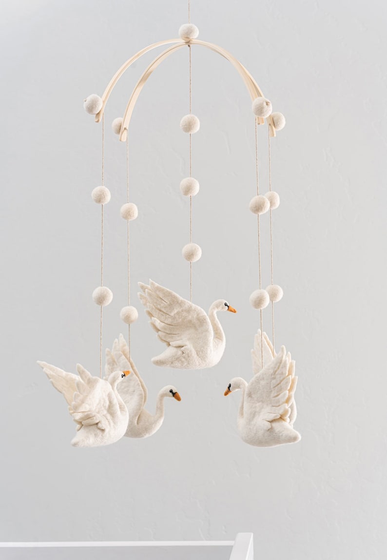 Puede incluir: Un m&oacute;vil de madera blanco con decoraciones de cisne de fieltro y pompones blancos colgando.