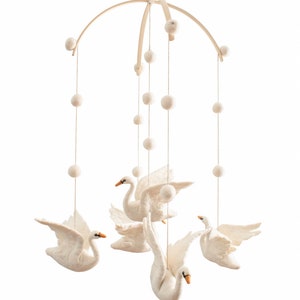 Puede incluir: Un m&oacute;vil de beb&eacute; de madera blanca con cinco cisnes de fieltro blanco y pompones blancos colgando del marco de madera.
