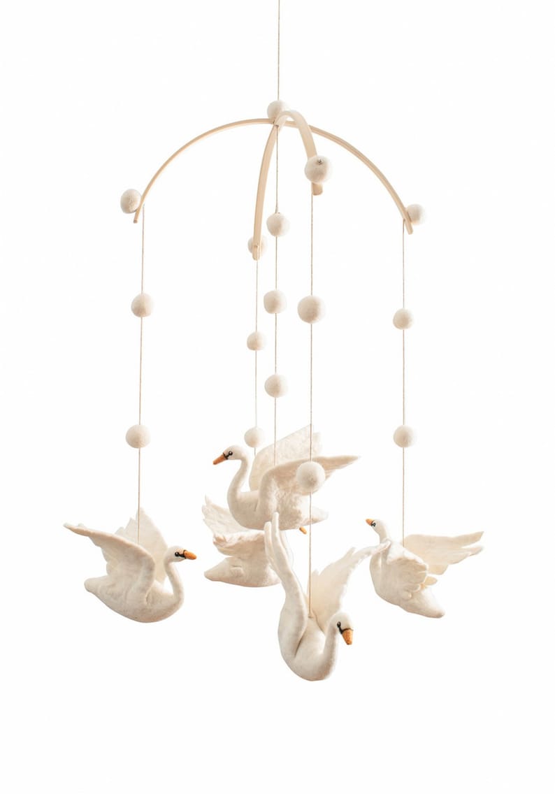 Puede incluir: Un m&oacute;vil de beb&eacute; de madera blanca con cinco cisnes de fieltro blanco y pompones blancos colgando del marco de madera.