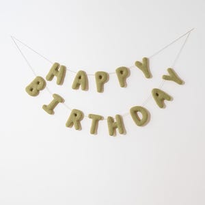 Può includere: Uno striscione in feltro verde con la scritta "HAPPY BIRTHDAY" in maiuscolo. Le lettere sono infilate su un filo sottile e trasparente su sfondo bianco. Lo striscione è una decorazione festosa per una festa di compleanno.