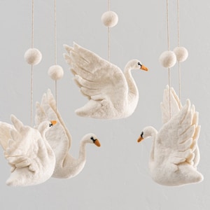 Puede incluir: Un m&oacute;vil de fieltro blanco con cuatro cisnes de fieltro blanco y cinco bolas de fieltro blanco colgando de un techo blanco.